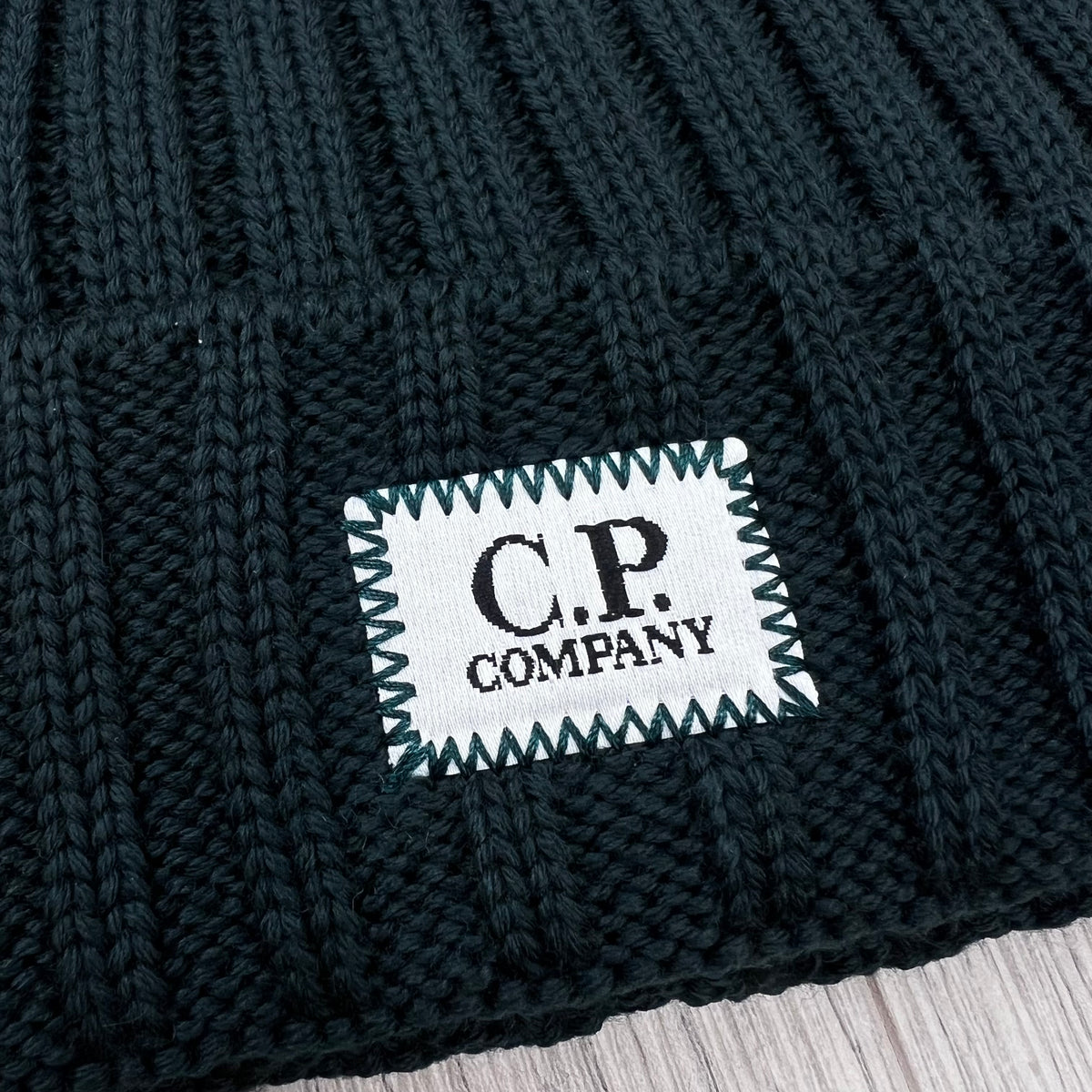 Close up view of the label logo embroidered onto a CP Company Merino Wool Patch Beanie in Ponderosa Pine .